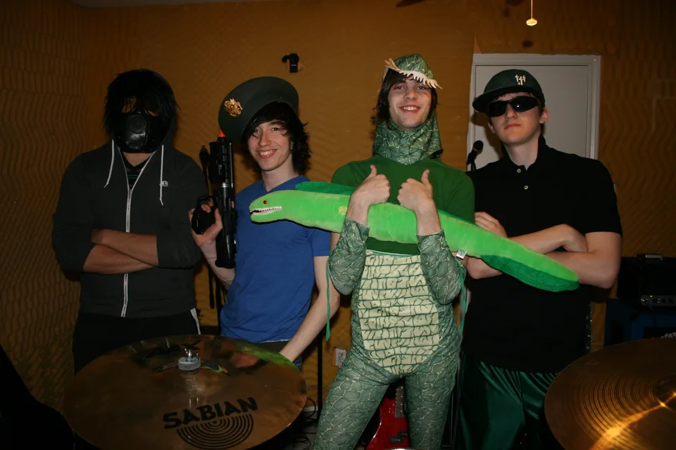 Four band members posing in costumes behind a drum kit, with a green stuffed dinosaur.