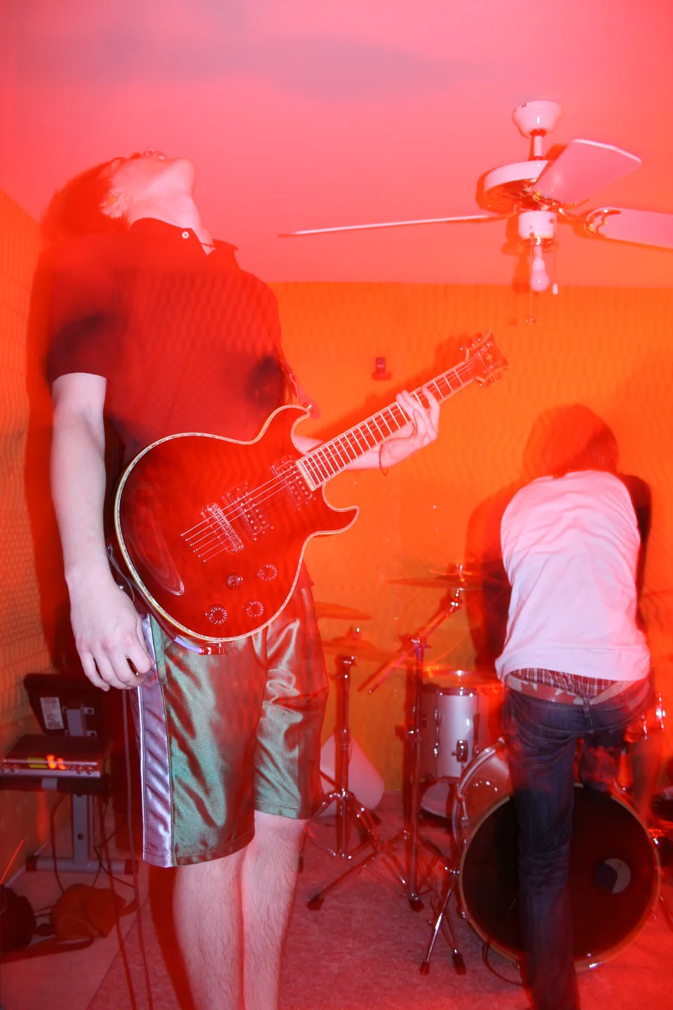 A guitarist leaning back beside a vocalist in an orange-lit practice room.