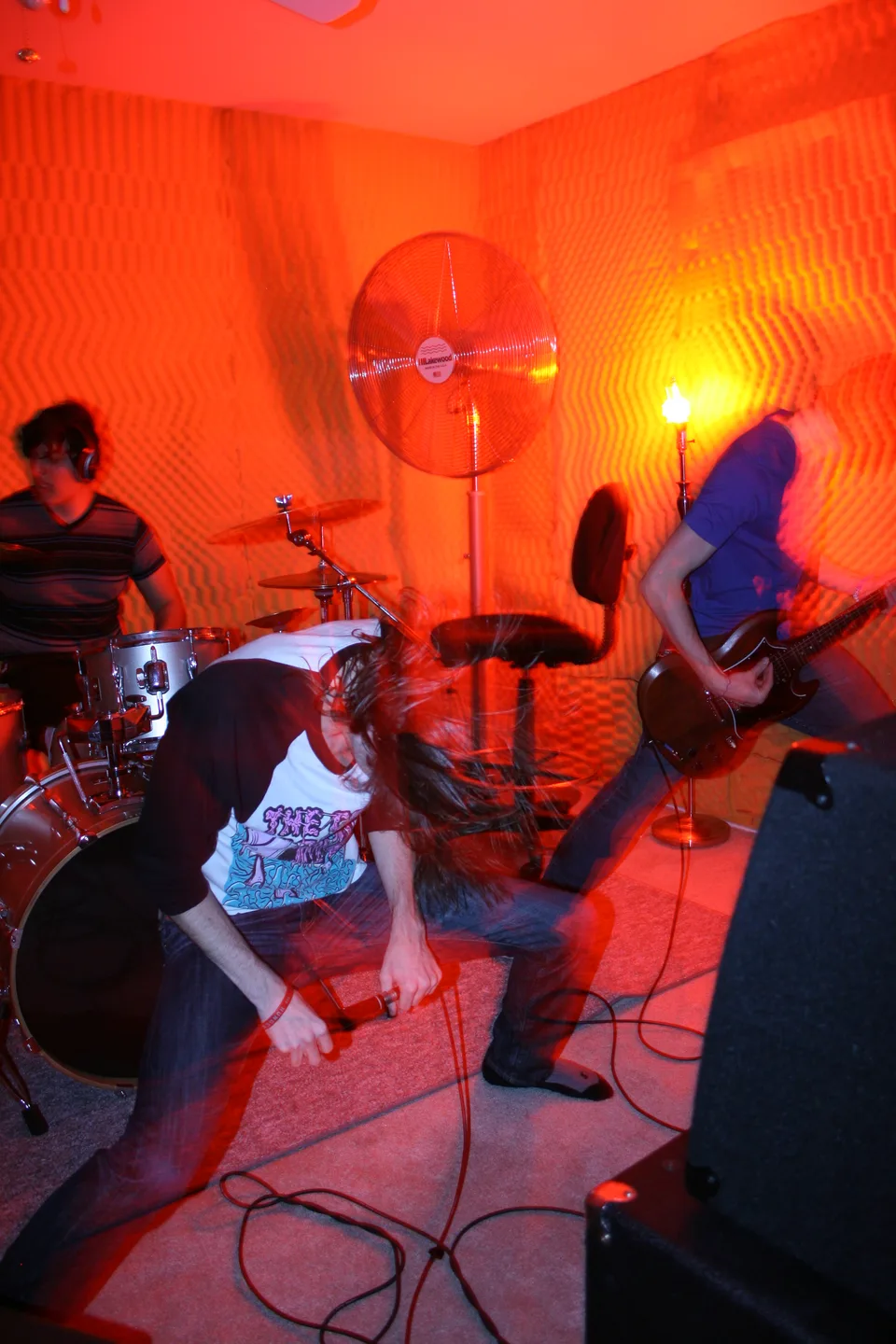 A vocalist crouching forward with a microphone, with a guitarist and drummer behind him.