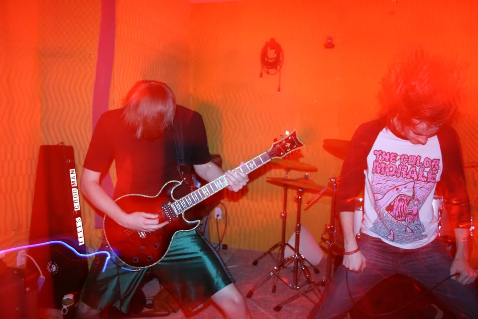 A guitarist and vocalist headbanging in front of a drum kit under orange light.
