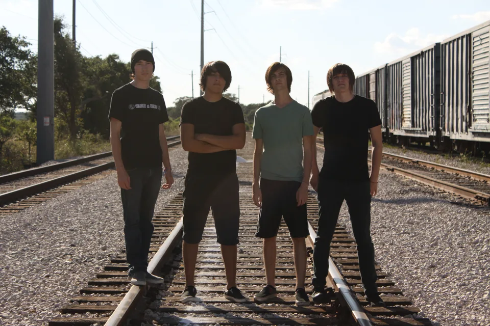 Four band members standing together on railway tracks beside freight cars.