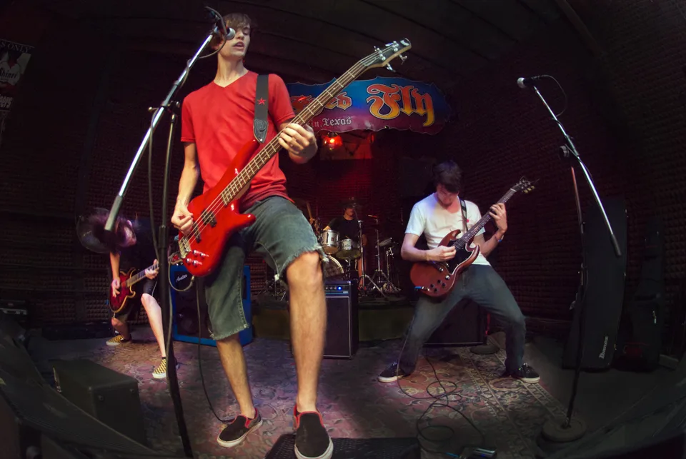 A bassist in a red shirt in the foreground, with the rest of the band performing behind him.